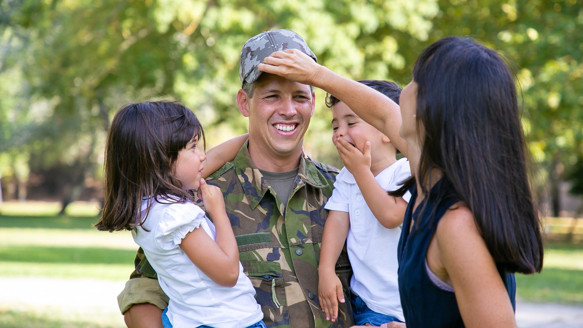 A smiling person in military uniform stands outdoors holding two children while another child reaches for their hat; all appear happy, showing moments of joy that help ease deployment depression.